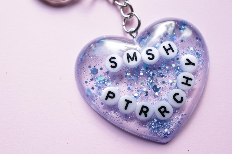 Może przedstawiać: Przezroczysty brelok w kształcie serca z niebieskimi błyskotkami i białymi literami tworzącymi napis "SMSHY PTRCHY".