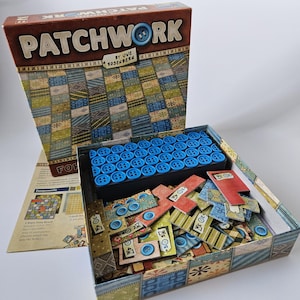 Peut inclure: Présentation du jeu de société "Patchwork", avec sa boîte et son contenu. La boîte est marron avec le titre du jeu et un bouton graphique. À l'intérieur, il y a des jetons bleus en forme de bouton, des pièces de jeu et un plateau de jeu. Les pièces de jeu ont diverses couleurs et motifs.