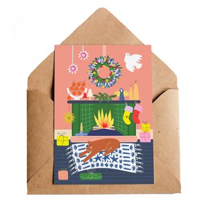 Puede incluir: Tarjeta de felicitaci&oacute;n navide&ntilde;a con una escena de chimenea y un gato marr&oacute;n durmiendo sobre una alfombra azul. La tarjeta incluye una corona, calcetines y regalos, todo sobre un fondo rosa, dentro de un sobre marr&oacute;n.