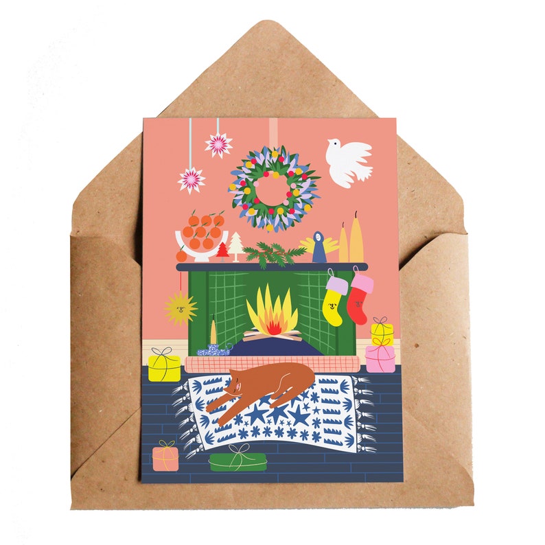 Puede incluir: Tarjeta de felicitaci&oacute;n navide&ntilde;a con una escena de chimenea y un gato marr&oacute;n durmiendo sobre una alfombra azul. La tarjeta incluye una corona, calcetines y regalos, todo sobre un fondo rosa, dentro de un sobre marr&oacute;n.