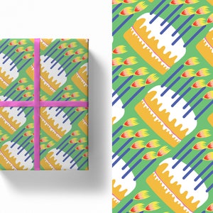 Puede incluir: Una caja de regalo envuelta en papel estampado con un diseño repetido de pasteles amarillos con glaseado blanco, líneas diagonales azules y formas de llama amarillas y rojas sobre un fondo verde. Una cinta rosa está atada alrededor de la caja.