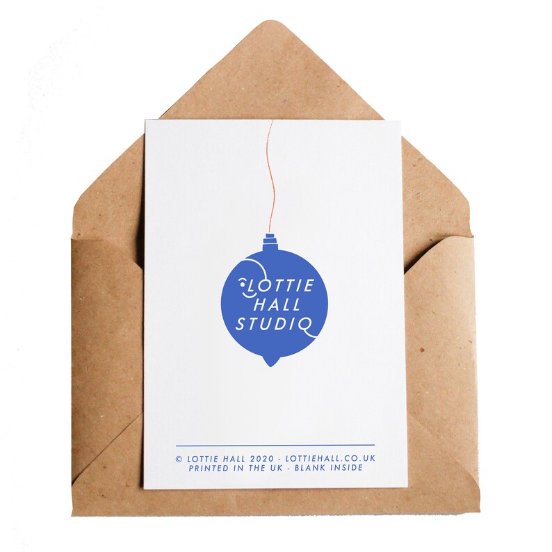 Puede incluir: Una tarjeta de felicitaci&oacute;n blanca con un dise&ntilde;o de adorno azul y el texto "Lottie Hall Studio" dentro de un sobre marr&oacute;n. La tarjeta tiene una cuerda roja unida al adorno. El sobre es de papel marr&oacute;n.