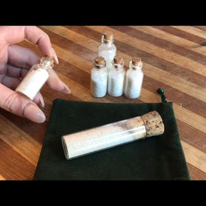May include: Six small glass bottles with cork stoppers filled with white crystals. The bottles are arranged in a row on a wooden surface. A larger glass bottle with a cork stopper is lying on a green velvet pouch.