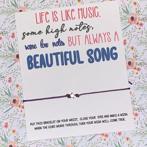 May include: A white card with the text "Life is like music, some high notes, some low notes but always a beautiful song."  A purple string bracelet with a silver star charm is attached to the card. The text "Put this bracelet on your wrist, close your eyes and make a wish. When the cord wears through, then your wish will come true." is printed on the card.