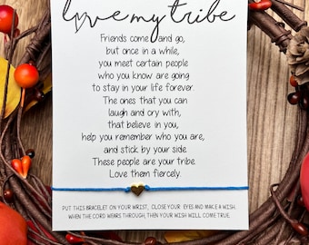 May include: A white card with the text "love my tribe" written in black cursive. The card has a quote about friendship and a blue string bracelet with a gold heart charm. The card is surrounded by a wreath of fall leaves.