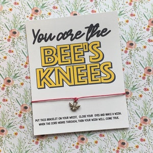 May include: A red string bracelet with a small bee charm. The card attached to the bracelet reads "You are the BEE'S KNEES" in yellow and black lettering. The card also includes instructions for making a wish when the string wears through.