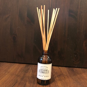 May include: A brown glass bottle with a white label that reads "Reed Diffuser" and "DICHENS CANDLES" with a white birch scent. The bottle is filled with a clear liquid and has wooden sticks inserted into it.