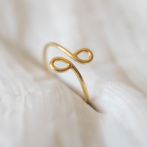 May include: A gold-toned ring with a simple, abstract design. The ring is made of a single piece of wire that is bent into two loops.