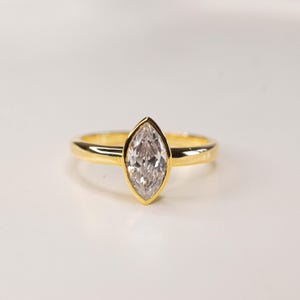 May include: A gold ring with a marquise-cut diamond. The diamond is set in a bezel setting, and the band is a simple, polished gold. The ring is displayed on a white surface.