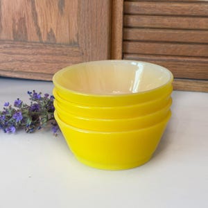 May include: A stack of four bright yellow glass bowls with a creamy white interior. The bowls are stacked neatly, showcasing their rounded shape. A sprig of purple flowers rests beside the bowls, adding a touch of color.