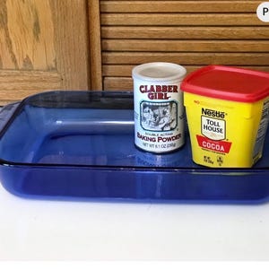 May include: A rectangular, cobalt blue glass baking dish with a black rim. Beside it are a can of Clabber Girl baking powder and a yellow container of Nestle Toll House cocoa with a red lid. The baking supplies are on a white surface.