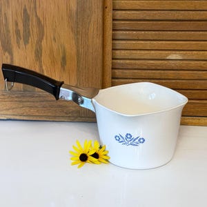 May include: White saucepan with a black handle and a blue floral design. The pan has a metal bracket and a small loop for hanging. Two yellow flowers with black centers are next to the pan.