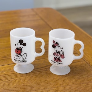 K&ouml;nnte beinhalten: Zwei wei&szlig;e Keramikbecher mit Mickey Mouse und Minnie Mouse. Jeder Becher hat einen einzigartigen Henkel und einen Sockel. Mickey ist in einer klassischen Pose, und Minnie h&auml;lt eine Handtasche. Die Becher stammen von Walt Disney Productions.