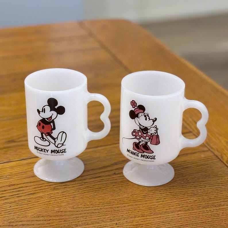 K&ouml;nnte beinhalten: Zwei wei&szlig;e Keramikbecher mit Mickey Mouse und Minnie Mouse. Jeder Becher hat einen einzigartigen Henkel und einen Sockel. Mickey ist in einer klassischen Pose, und Minnie h&auml;lt eine Handtasche. Die Becher stammen von Walt Disney Productions.