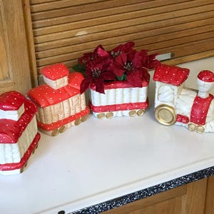 May include: A collection of ceramic train figurines in red and white, with red polka dots. One train car holds red flowers. The train cars are arranged on a white surface, suggesting a decorative theme.