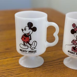 K&ouml;nnte beinhalten: Zwei wei&szlig;e Tassen mit Fu&szlig; und einem Mickey Mouse und Minnie Mouse Design. Die Mickey Mouse Tasse zeigt eine schwarz-rote Grafik von Mickey Mouse mit dem Text "Mickey Mouse" und "Walt Disney Productions". Die Minnie Mouse Tasse ist ebenfalls sichtbar.