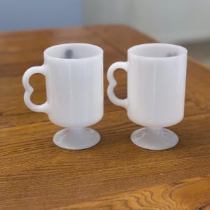 K&ouml;nnte beinhalten: Zwei wei&szlig;e Tassen im Vintage-Stil mit herzf&ouml;rmigen Griffen und Sockeln. Die Tassen bestehen aus undurchsichtigem Glas und stehen auf einer Holzoberfl&auml;che. Die Tassen sind leer.