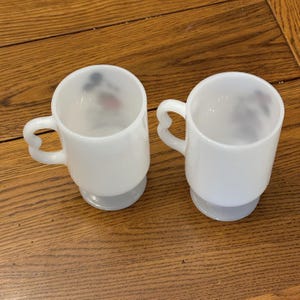 K&ouml;nnte beinhalten: Zwei wei&szlig;e Glasbecher mit herzf&ouml;rmigen Griffen. Die Becher haben einen abgerundeten Boden und einen leicht ausgestellten Rand. Im Inneren der Becher ist ein schwaches Bild einer Zeichentrickfigur zu sehen. Die Becher stehen auf einer Holzoberfl&auml;che.