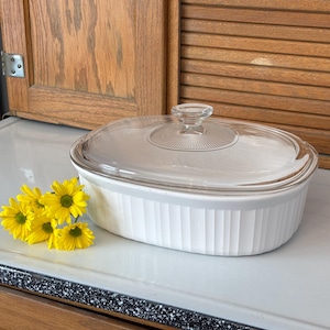 May include: White oval casserole dish with a clear glass lid. The dish has vertical ribbed detailing and is next to a small bouquet of yellow flowers. The dish is on a white surface.
