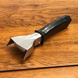 May include: A black and silver pot handle. The handle is black and has a geometric design. The part that attaches to the pot is silver and has a curved design. The handle is lying on a wooden surface.