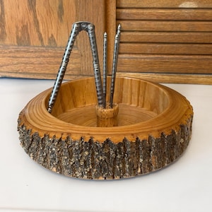 May include: A wooden nutcracker set featuring a round bowl with a natural bark edge. The set includes a metal nutcracker and two picks. The bowl is made of light-colored wood, and the nutcracker tools are silver.
