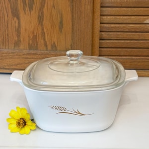 May include: White square casserole dish with a clear glass lid and a wheat design. The dish has two handles and a clear knob on the lid. A yellow flower is next to the dish.