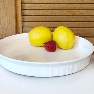 May include: A white, round, ceramic pie dish with a fluted edge. Two yellow lemons and three red strawberries are arranged on the dish. The background includes wooden shutters and a white surface.