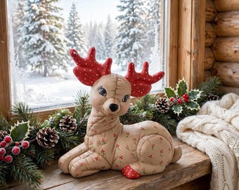 May include: A tan fabric deer figurine with a stitched texture, featuring large red antlers with white polka dots and a small red berry pattern on its body. The deer has long eyelashes, a black nose, and is in a seated, resting position.