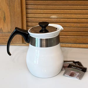 May include: White ceramic teapot with a black handle and lid. A silver band encircles the middle of the teapot. Three Earl Grey tea packets are next to the teapot. The teapot is on a white surface.