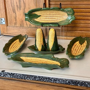 May include: A ceramic corn-themed kitchen set. The set includes two salt and pepper shakers, three serving dishes, and a decorative tray. The pieces are green and yellow, with a detailed corn cob design.