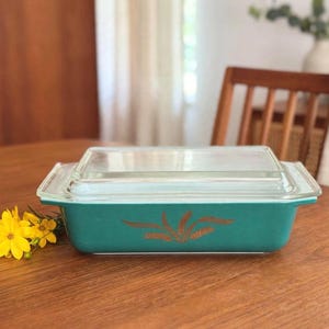 May include: A rectangular teal glass casserole dish with a clear glass lid. The exterior of the dish features a gold-colored wheat stalk pattern centered on the side. The dish has integrated handles on both ends and a smooth, glossy finish.