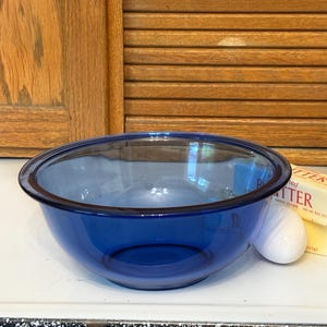 May include: A clear, cobalt blue glass mixing bowl. The bowl is round with a slightly flared rim. A stick of butter and an egg are next to the bowl. The background includes wooden shutters and a wooden door.