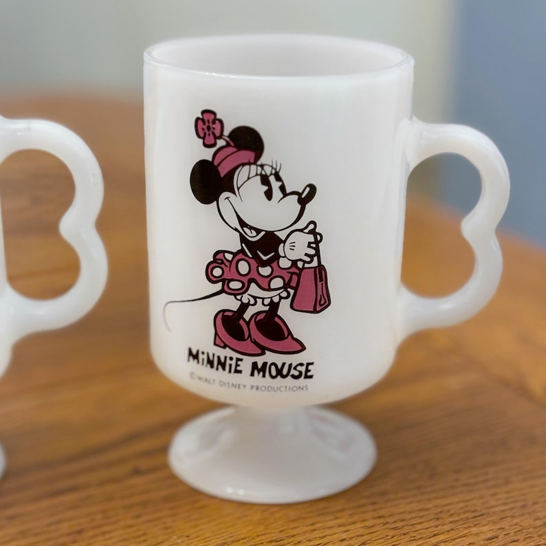 K&ouml;nnte beinhalten: Wei&szlig;er Minnie Mouse-Becher aus Glas mit herzf&ouml;rmigem Griff und Sockel. Der Becher zeigt eine Minnie Mouse-Grafik in Schwarz, Rot und Rosa mit dem Aufdruck "MINNIE MOUSE" darunter. &copy; Walt Disney Productions.