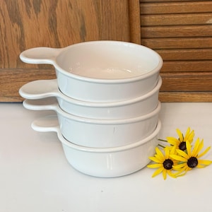 May include: A stack of four white ceramic bowls, each featuring a single small handle on the side. The bowls have a smooth, glossy finish and a rounded shape. The stack is positioned next to three yellow and brown coneflower blossoms.