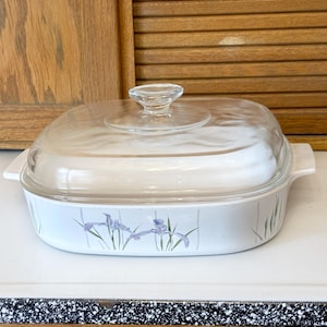 May include: Square, off-white casserole dish with a clear glass lid. The dish features a light blue floral design with purple irises and green stems. The dish has two white handles.