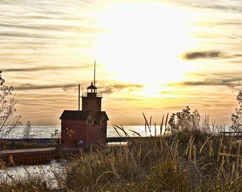 May include: A red lighthouse stands tall against a golden sunset sky. The lighthouse is located on a small peninsula with a grassy area in the foreground. The water is calm and the sky is clear.