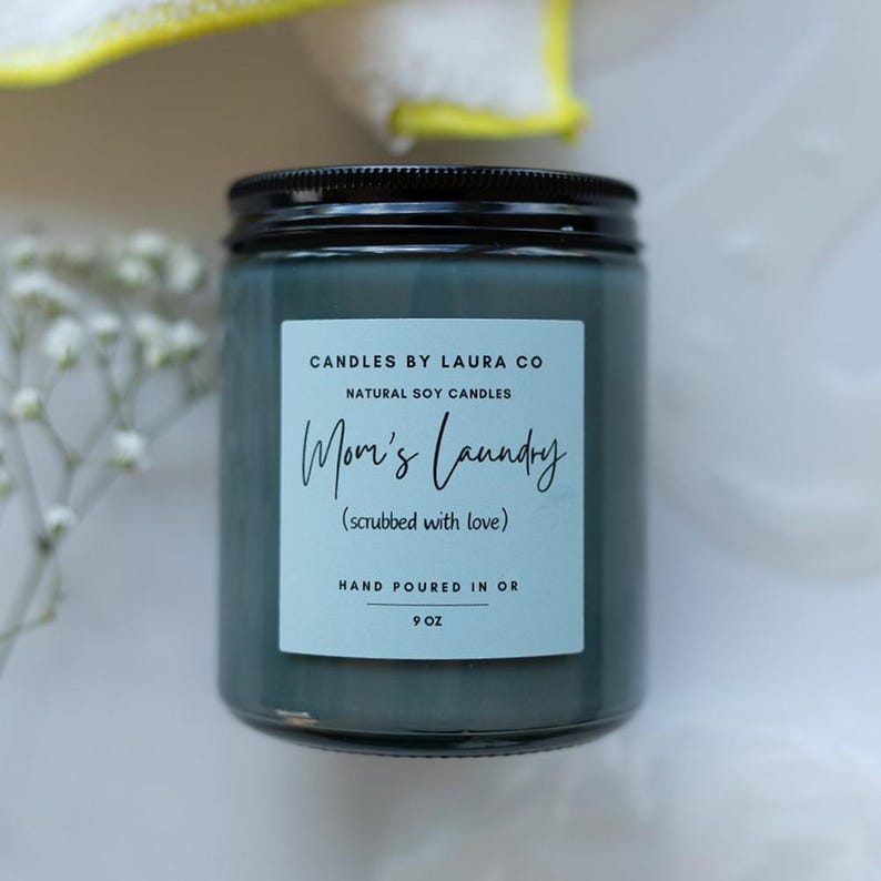 May include: A teal blue glass candle jar with a black lid. The label reads "CANDLES BY LAURA CO NATURAL SOY CANDLES Mom's Laundry (scrubbed with love) HAND POURED IN OR 9 OZ".