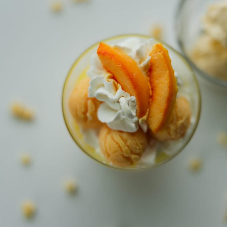 Puede incluir: Un postre en vaso con bolas de sorbete de melocot&oacute;n naranja, cubierto con nata montada blanca y dos rodajas de melocot&oacute;n fresco. El postre est&aacute; dispuesto en un cuenco de cristal transparente, mostrando la fruta naranja y la crema blanca.