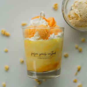 Puede incluir: Una vela de cristal transparente rellena de cera amarilla, cubierta con cera batida blanca y bolas de cera naranja. La etiqueta dice: frozen peach custard, AND POURED BY CANDLES BY LAURA, SOYBEAN WAX/NET WT 12 OZ. Un cuenco de helado est&aacute; al lado.