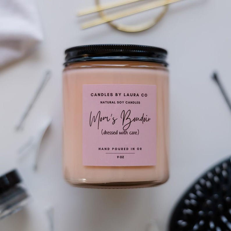 May include: A pink and white candle jar with a black lid. The label reads "CANDLES BY LAURA CO NATURAL SOY CANDLES Mom's Boudoir (dressed with care) HAND POURED IN OR 9 OZ".