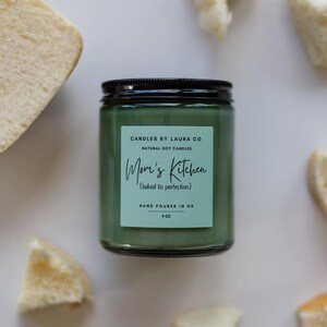 May include: A green glass candle jar with a black lid. The label reads "CANDLES BY LAURA CO NATURAL SOY CANDLES Mom's Kitchen (baked to perfection) HAND POURED IN OR 9 OZ".