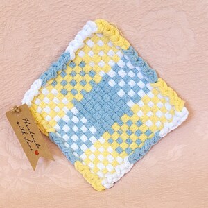 May include: A square woven pot holder with a blue, white, and yellow checkered pattern. The pot holder has a braided border and a brown tag that says "Handmade with love".
