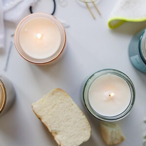 May include: Four candles in glass jars with a white wax and a small flame burning in each. The candles are on a white surface with a piece of bread and some small white flowers.