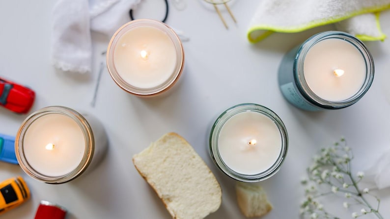 May include: Four candles in glass jars with a white wax and a small flame burning in each. The candles are on a white surface with a piece of bread and some small white flowers.