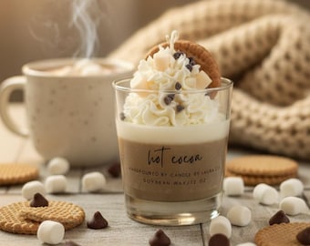May include: A clear glass candle filled with brown wax, topped with white wax whipped cream, chocolate chips, and a cookie. The label reads hot cocoa, handpoured by candle by laura co, soybean wax/12 oz. The candle is surrounded by marshmallows and cookies.