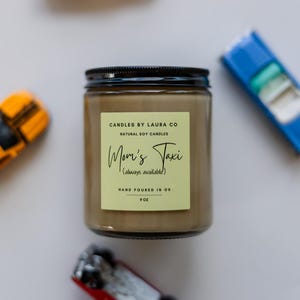 May include: A brown glass candle jar with a black lid. The label is light green with black text that reads "CANDLES BY LAURA CO NATURAL SOY CANDLES Mom's Taxi (always available) HAND POURED IN OR 9 OZ".