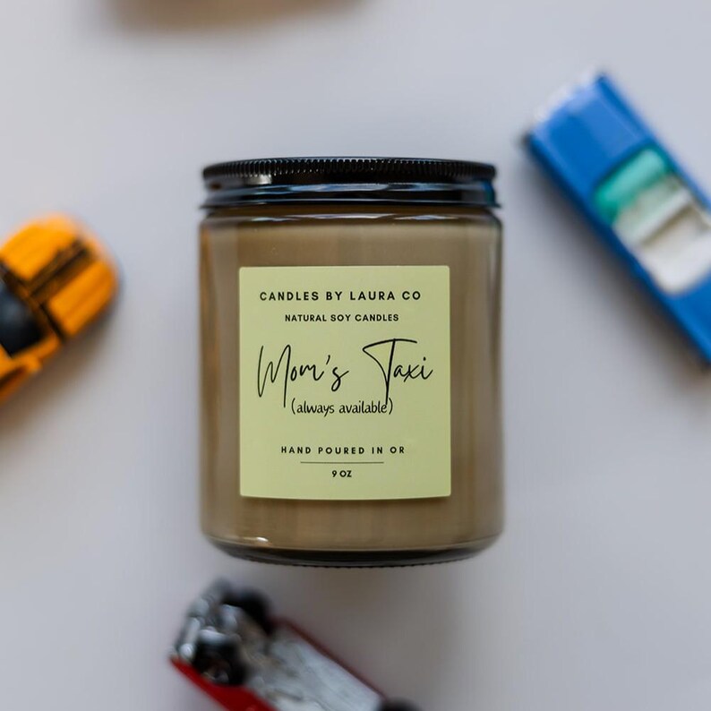 May include: A brown glass candle jar with a black lid. The label is light green with black text that reads "CANDLES BY LAURA CO NATURAL SOY CANDLES Mom's Taxi (always available) HAND POURED IN OR 9 OZ".