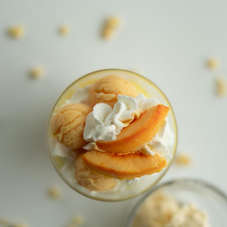 Puede incluir: Un vaso lleno de un postre que presenta bolas de sorbete o helado de color naranja, cubierto con un generoso remolino de nata montada blanca y dos rodajas de melocot&oacute;n fresco. El postre se presenta desde una perspectiva cenital.