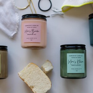May include: Four candles with black lids and labels that say "Mom's" followed by a different word. The candles are in shades of pink, brown, green, and blue. The candles are on a white surface with other items, including toy cars, bread, and a white cloth.