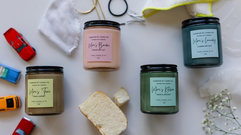 May include: Four candles with black lids and labels that say "Mom's" followed by a different word. The candles are in shades of pink, brown, green, and blue. The candles are on a white surface with other items, including toy cars, bread, and a white cloth.